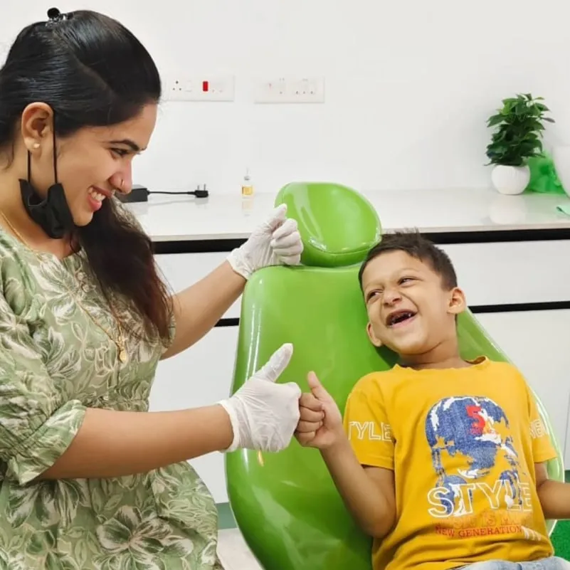 A dentist and a laughing young boy sharing a thumbs-up beside the green dental chair